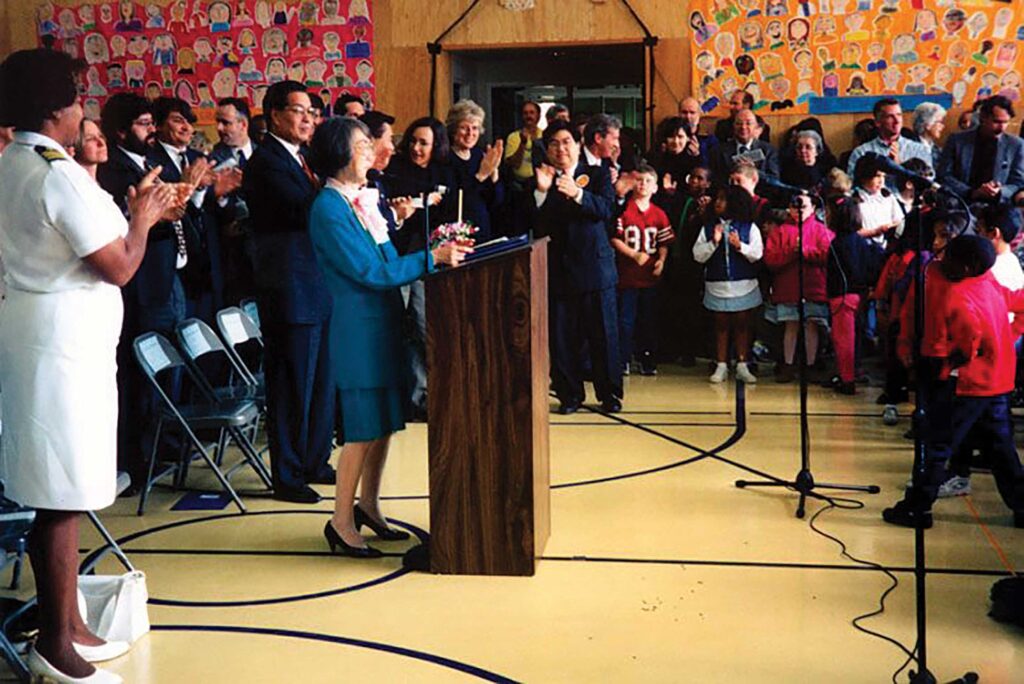 Honored guests, including SFUSD Superintendent Bill Rojas, stand applauding as principal Nancy Mayeda stands at the speaker podium in the school gym for the dedication of the Nancy Yoshihara Mayeda Campus.