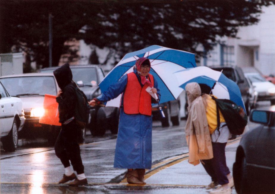 Principal Nancy Mayeda, dressed in a blue raincoat and orange safety vest, stops traffic under a big blue and white umbrella to help students cross the street.